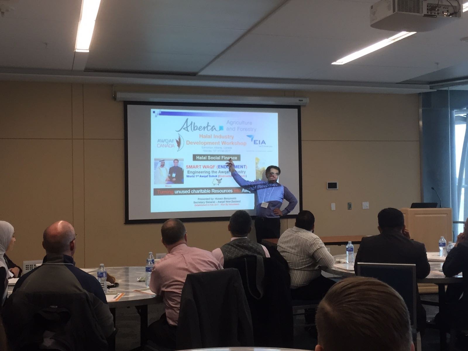 Husain Benyounis presents in front of a projected title slide reading Halal Industry Development Workshop, Halal Social Finance, Smart Waqf, presented by Husain Benyounis, Secretary General, Awqaf New Zealand, while participants seated at round tables listen, seen from behind.